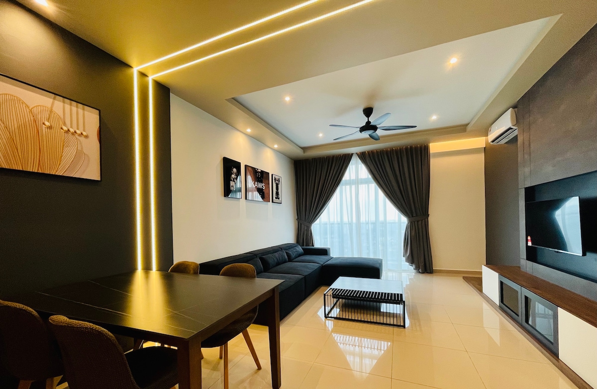 A modern living room area features a dark sectional sofa positioned beside a contemporary coffee table. A dining table with four chairs is present. Large windows with flowing curtains allow natural light to brighten the space, complemented by wall-mounted artwork and ceiling lighting.