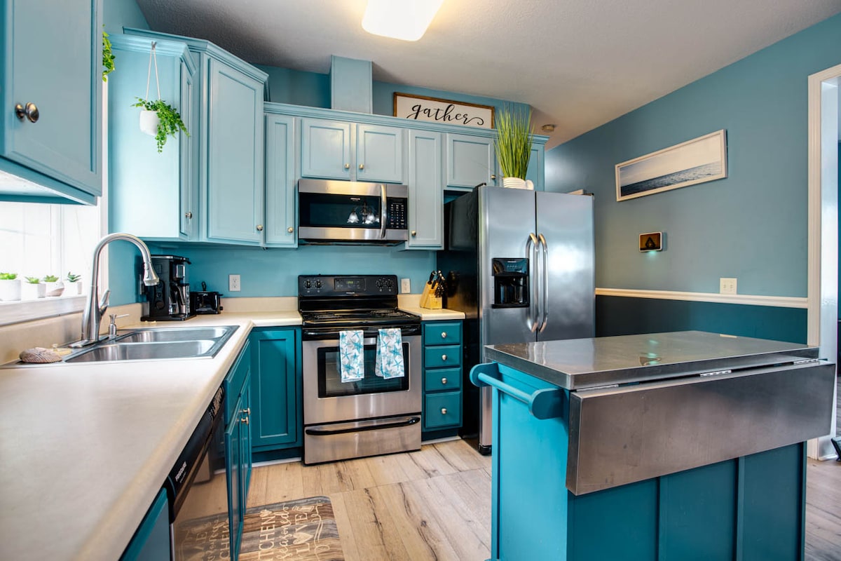 A modern kitchen showcases blue cabinetry and stainless steel appliances. A spacious center island offers additional counter space, with a sink and storage drawers integrated into the design. Natural light enters through a nearby window, enhancing the fresh atmosphere.