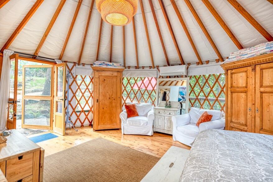 The interior of the yurt features a circular layout with natural wood accents. Two white upholstered chairs are positioned beside a decorative cabinet. An ornate lighting fixture hangs from the ceiling, while the entrance door opens to the outside, inviting natural light into the cozy space.