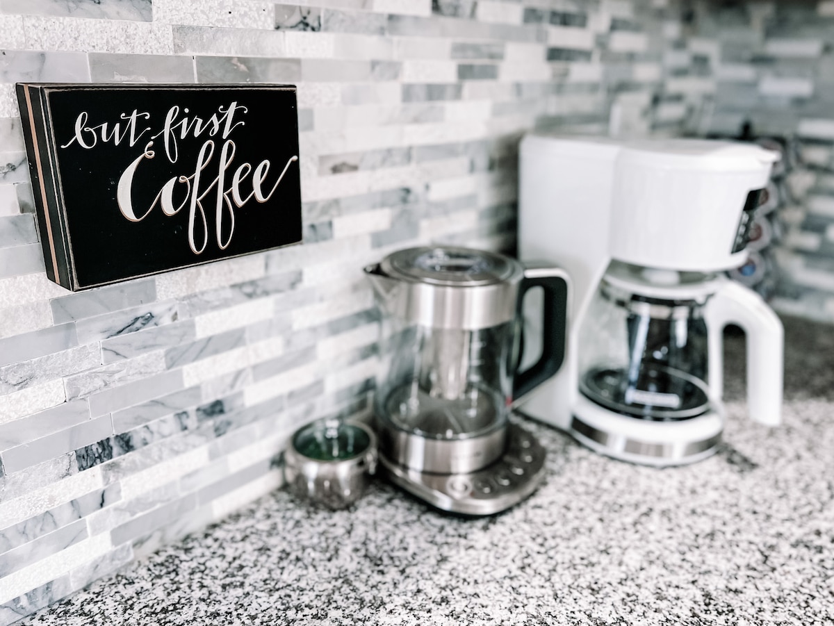 A modern kitchen countertop is displayed, featuring a coffee maker and an electric kettle in stainless steel. A decorative sign with the phrase 'but first coffee' is positioned above, against a backdrop of textured stone tiles. The surface is composed of a speckled gray material.