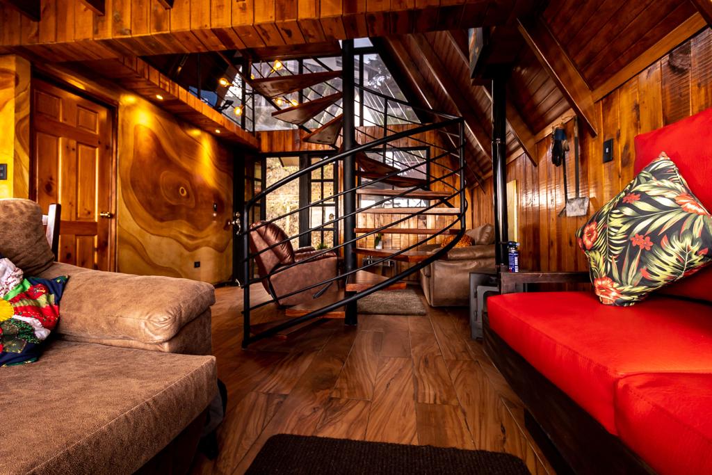 A warm interior space features wooden walls and a spiral staircase as a central element. Comfortable seating options include a light brown sofa and a vibrant red couch adorned with decorative pillows. Soft lighting enhances the cozy ambiance throughout the room.