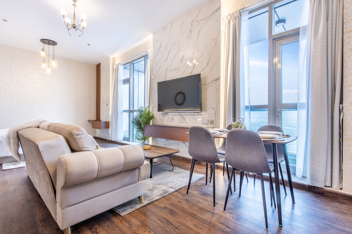 A modern living area features a comfortable seating arrangement with a light-colored sofa and a wooden coffee table. A flat-screen television is mounted on a marble accent wall, while large windows allow natural light to fill the space, complemented by soft curtains.