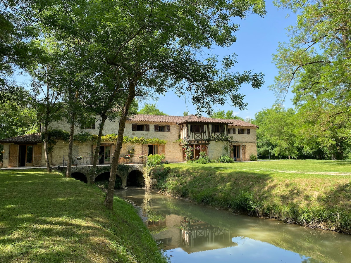 The exterior of the restored moulin is nestled among lush greenery, with a serene waterway reflecting the building's charming architecture. The two-story structure features a combination of stone and wooden elements, while large windows offer views of the surrounding nature.