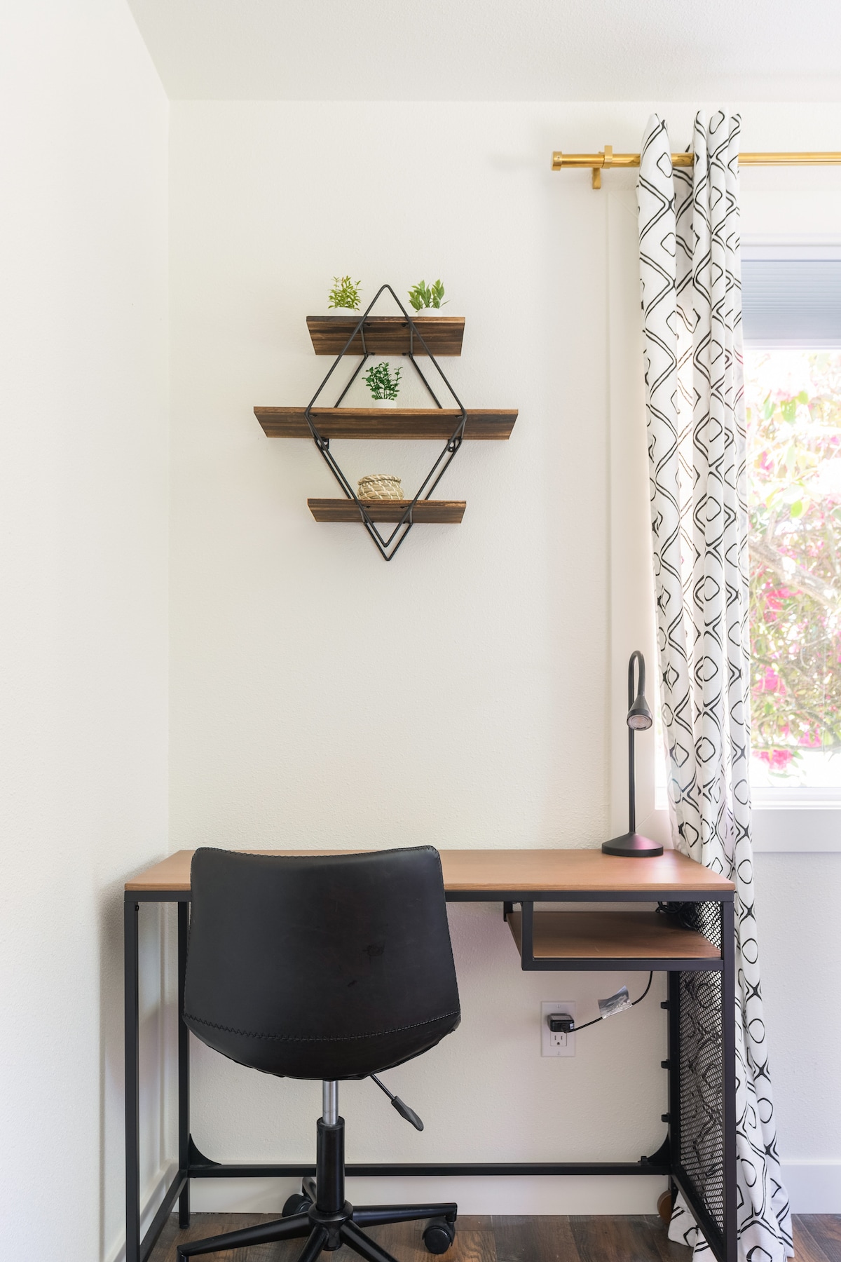 A modern workspace is presented with a sleek desk and a rolling chair. Above the desk, three wooden shelves display small potted plants, while a lamp provides task lighting. A window, partially adorned with patterned drapes, allows natural light to brighten the area.