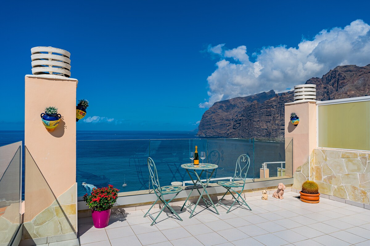 A spacious terrace is visible, featuring a small glass table and two metal chairs. Colorful flower pots decorate the walls, and a scenic view of the ocean and cliffs is framed against a bright blue sky with scattered clouds.