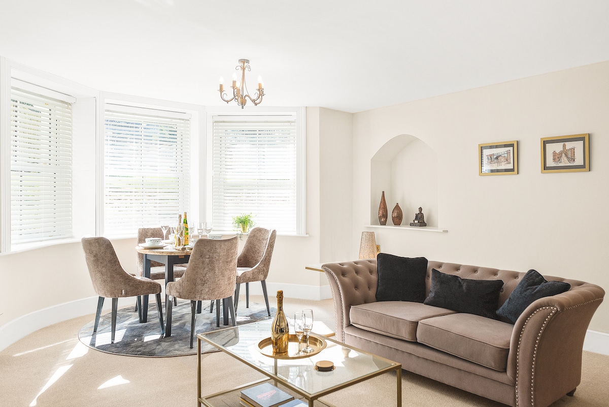 A bright living area is observed, featuring a comfortable upholstered sofa and a round dining table surrounded by plush chairs. Natural light filters through multiple windows, enhancing the elegant decor. A decorative chandelier adds a subtle touch above the seating arrangement.