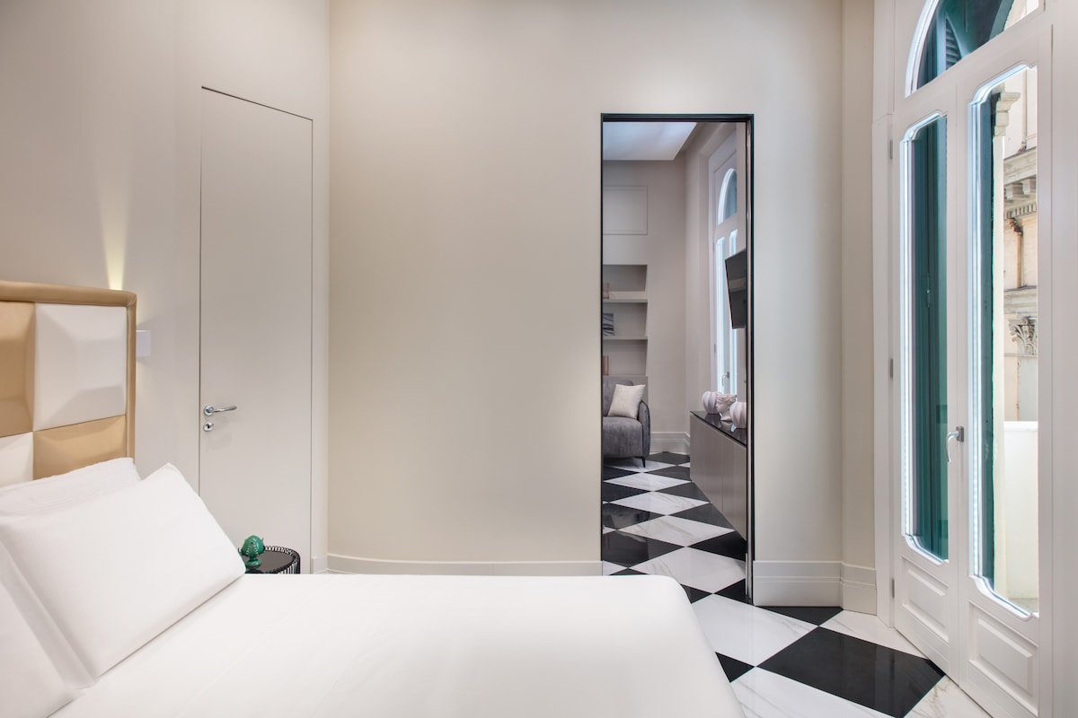 A spacious bedroom features a large bed dressed in crisp white linens. A doorway with a mirror reflects the adjacent sitting area. Tall windows with shutters allow natural light to fill the room, while geometric black and white flooring adds a modern touch.
