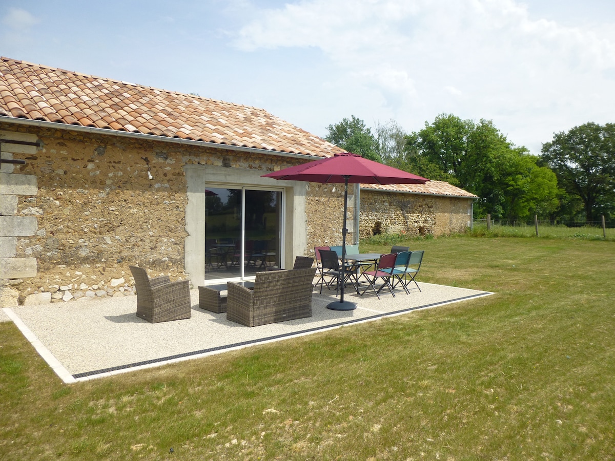 A terrace area features a set of colorful chairs and a table under a large umbrella. Comfortable seating arrangements are positioned nearby on gravel, surrounded by a grassy field and trees in the background. The stone walls of the barn are visible, complementing the outdoor space.