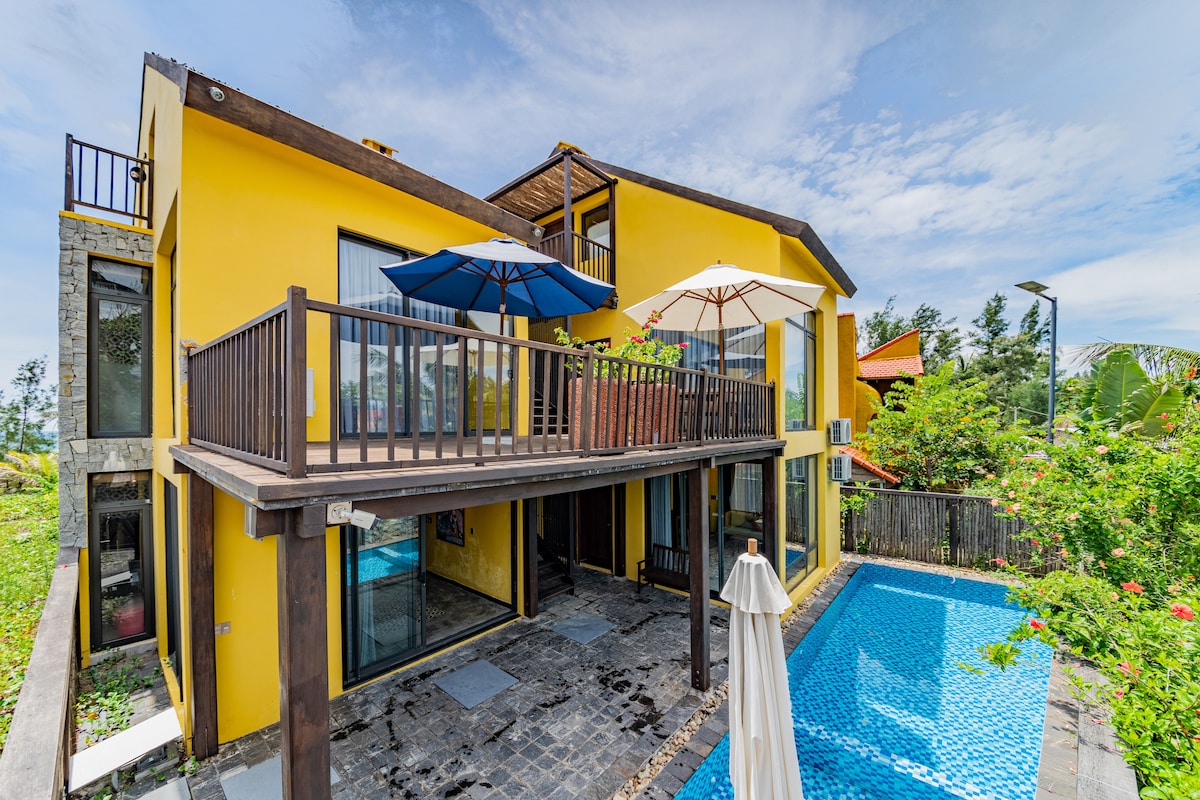 The exterior of the villa is showcased, featuring a modern yellow facade with large glass windows. A private pool is visible in the foreground, complemented by a stone patio. A balcony with umbrellas provides seating space, surrounded by tropical greenery.
