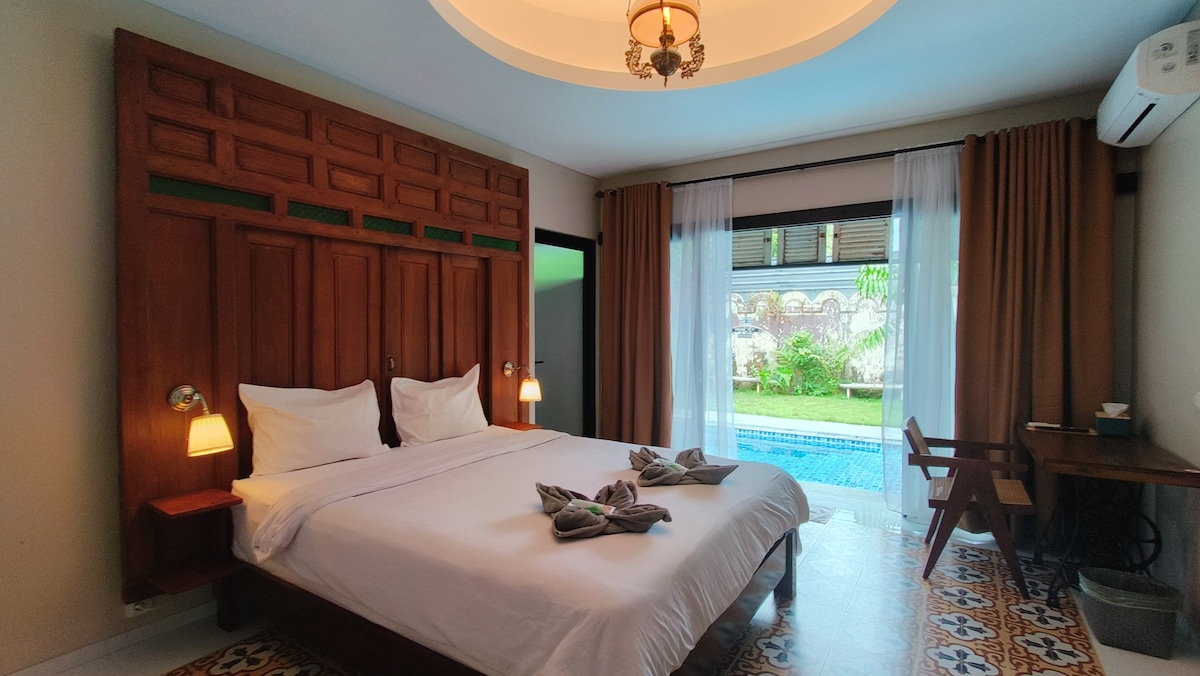 A spacious room features a wooden headboard with traditional carvings and a large bed. Soft lighting is provided by bedside lamps. Large windows overlook a garden and swimming pool, with curtains allowing natural light to filter through. A wooden desk is positioned beside the window.
