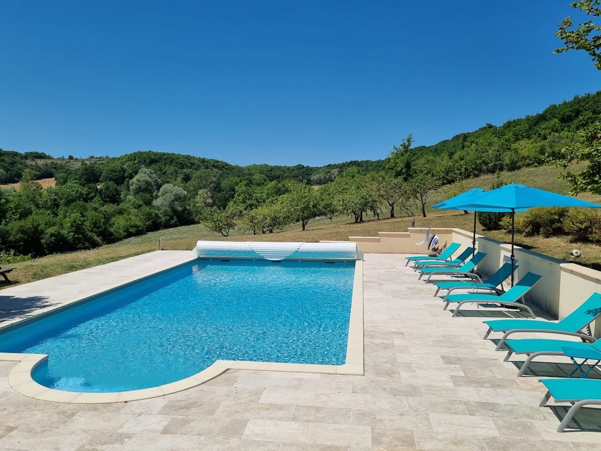 A heated swimming pool is framed by a spacious terrace, featuring turquoise loungers and umbrellas. Lush greenery and rolling hills are visible in the background, creating a serene natural environment for relaxation.