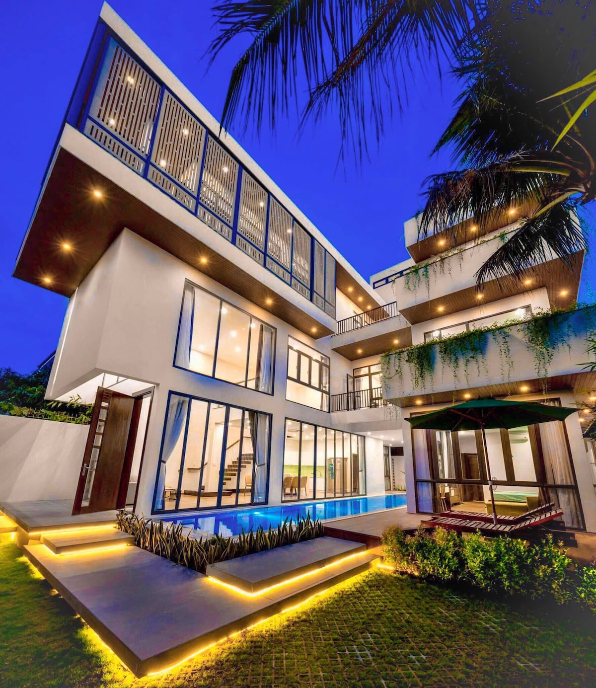 The villa's modern architecture is showcased at dusk, with large glass windows reflecting warm indoor lighting. A long swimming pool runs alongside the building, surrounded by lush greenery and decorative lighting that enhances the outdoor space.
