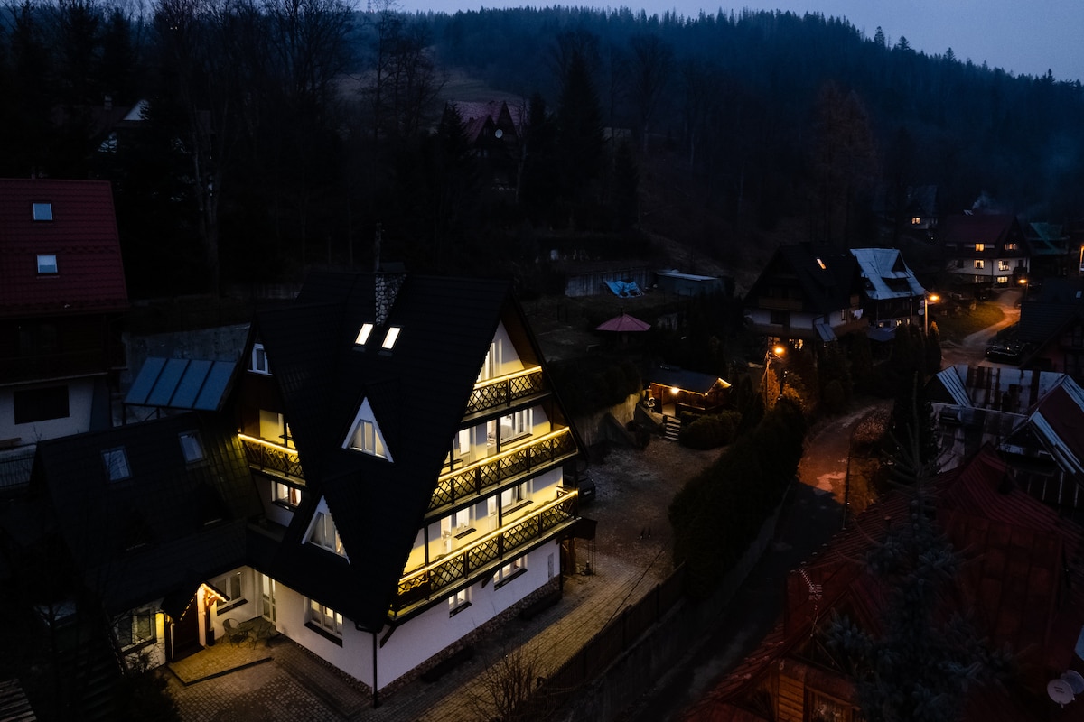 A charming multi-story building is illuminated softly at dusk, showcasing its architectural details. Surrounding greenery and an inviting path lead to the entrance. Nearby homes are nestled within a tranquil setting, providing a sense of harmony with the natural landscape.