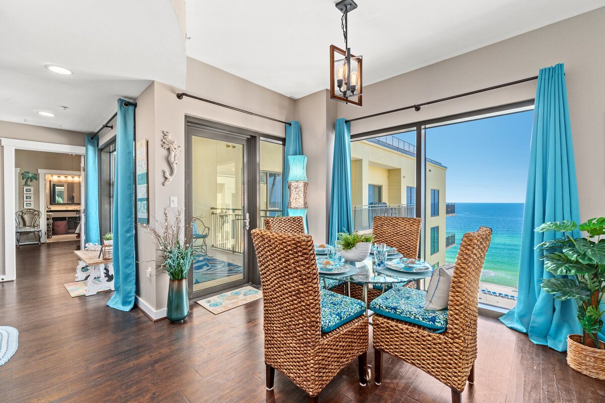 A dining area features a glass table surrounded by woven chairs, adorned with blue accents. Floor-to-ceiling windows offer a panoramic view of the ocean, while vibrant blue curtains frame the scenic backdrop. A coastal theme is evident in the decor, enhancing the dining experience.