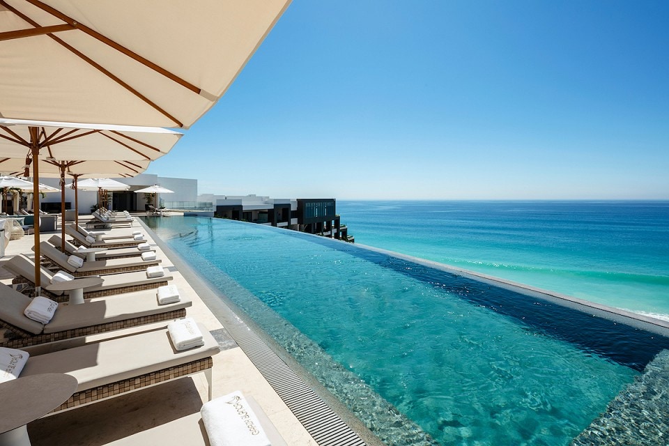 An infinity pool extends toward the horizon, merging with the bright blue ocean. Sun loungers are positioned under umbrellas along the pool's edge, while the clear water reflects the surrounding sky. The distant coastline is visible, enhancing the serene setting.