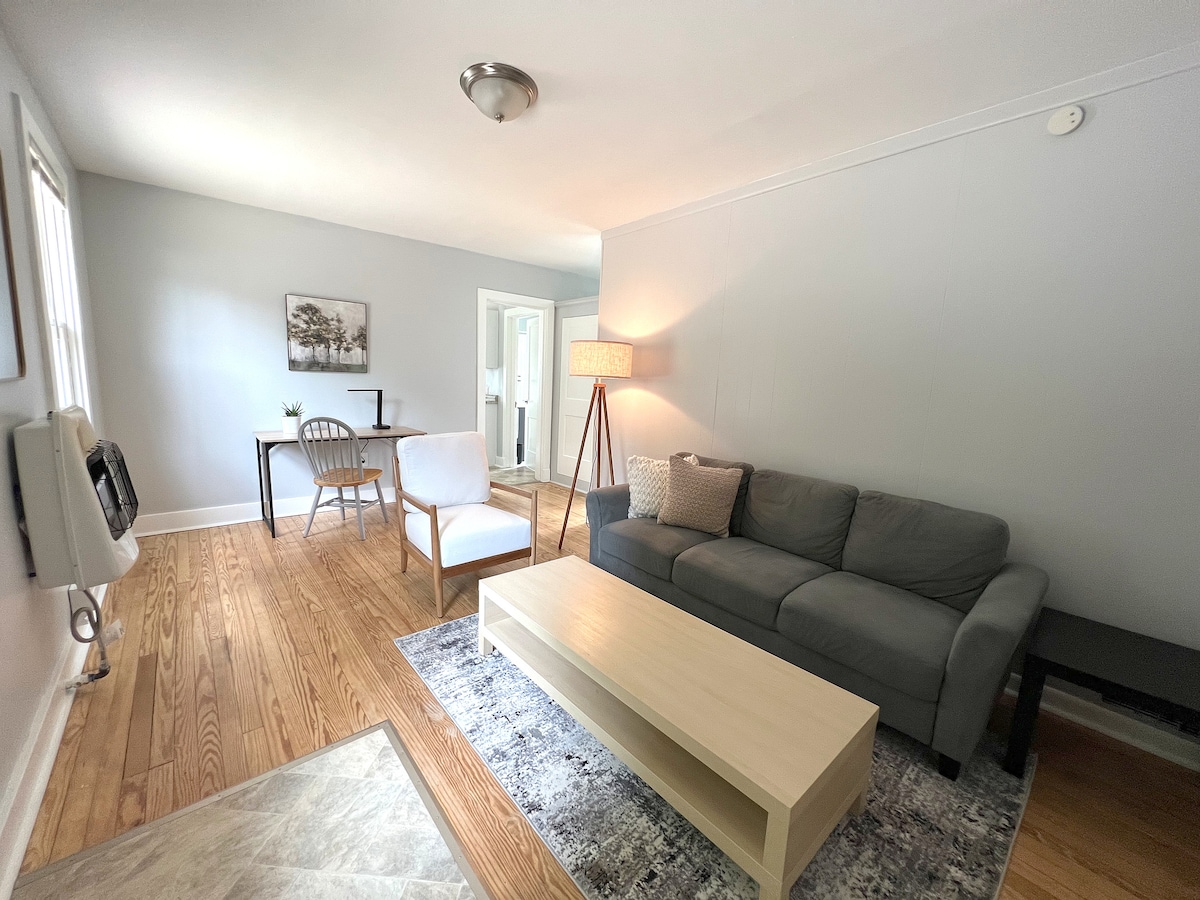 A spacious living area is highlighted by hardwood flooring and neutral-colored walls. A soft gray sofa is positioned across from a light-colored coffee table, with a cozy armchair nearby. Natural light filters through the windows, enhancing the inviting ambiance.