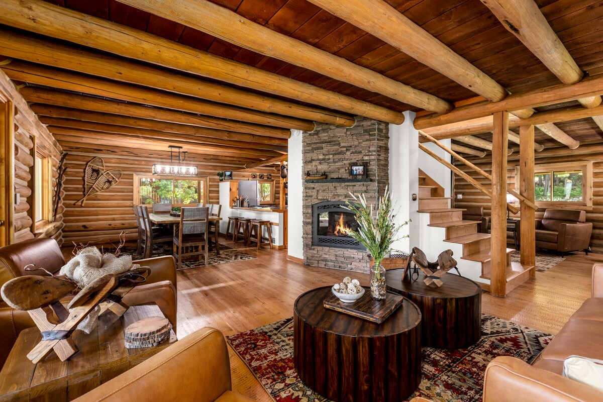 The main living area features a stone fireplace surrounded by cozy leather seating and wooden accents. A large dining area is visible in the background, illuminated by natural light from nearby windows. A rustic coffee table adds warmth to the inviting atmosphere of the space.