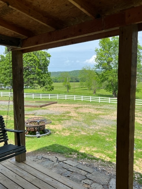 A view from the porch reveals a serene landscape of green fields bordered by a white fence. A gravel area is visible, alongside a circular fire pit with seating. The scene is filled with trees and distant hills under a clear sky.