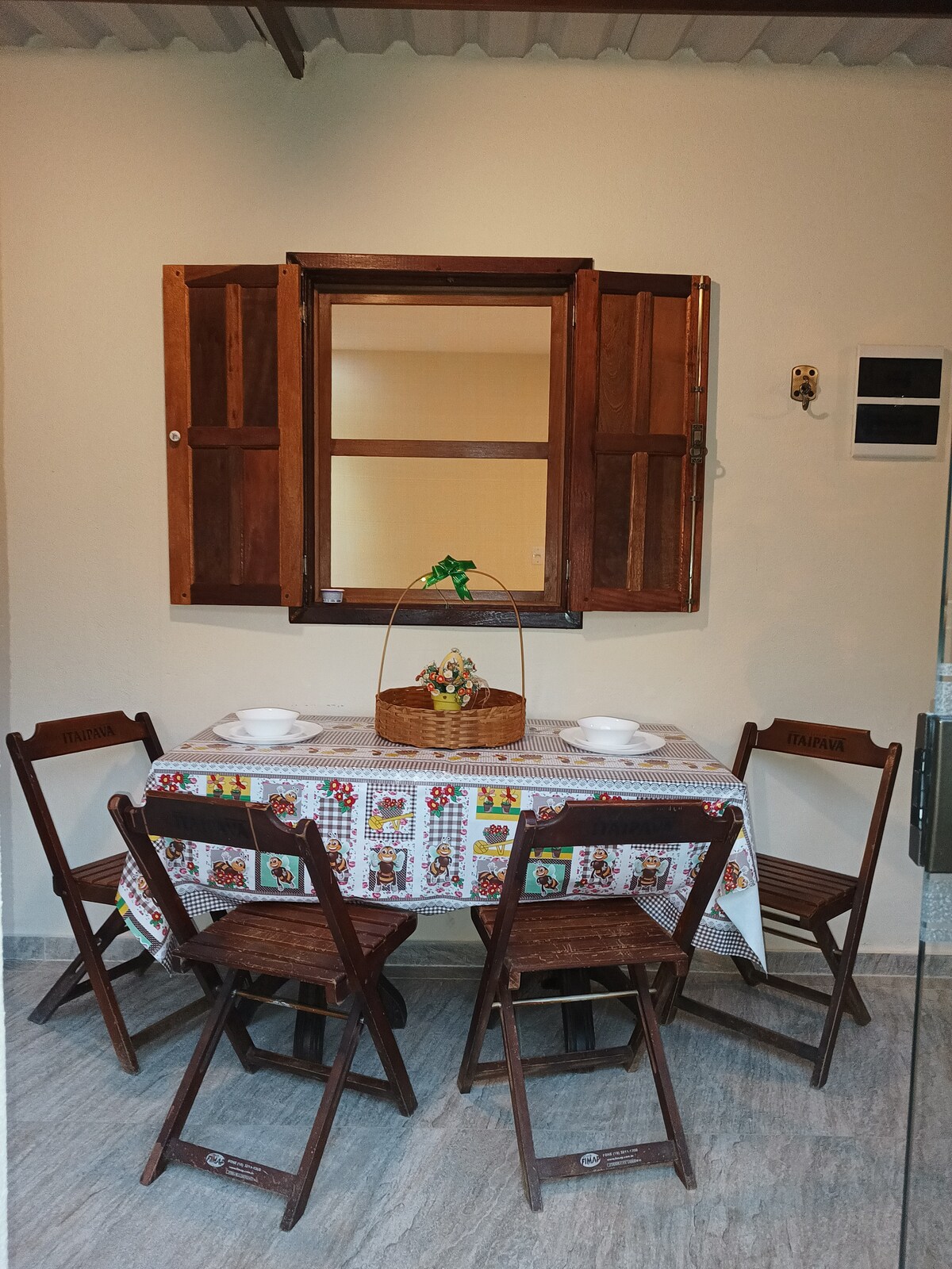 A quaint dining area is furnished with a small table covered by a patterned tablecloth. Four wooden chairs are arranged around the table, which holds a decorative basket at its center. Wooden shutters frame a window that allows natural light to enter the space.
