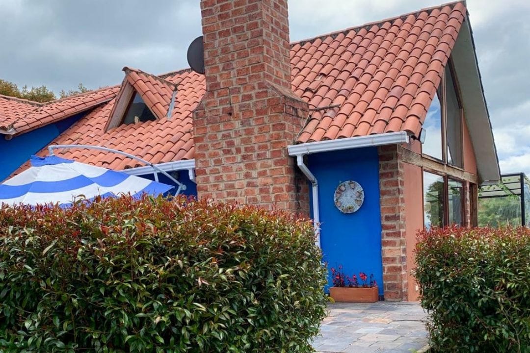 The exterior of the country home features a vibrant blue wall complemented by a red tile roof. A brick chimney is visible, while lush greenery forms a border along the pathway. A round clock adorns the wall, adding a subtle charm to the outdoor space.