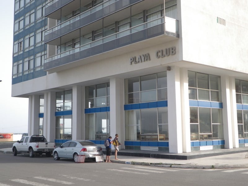 The building features a modern facade with large glass windows and a prominent sign reading 'Playa Club.' The entrance is easily accessible, and vehicles are seen parked in front, illustrating the building's convenient location.