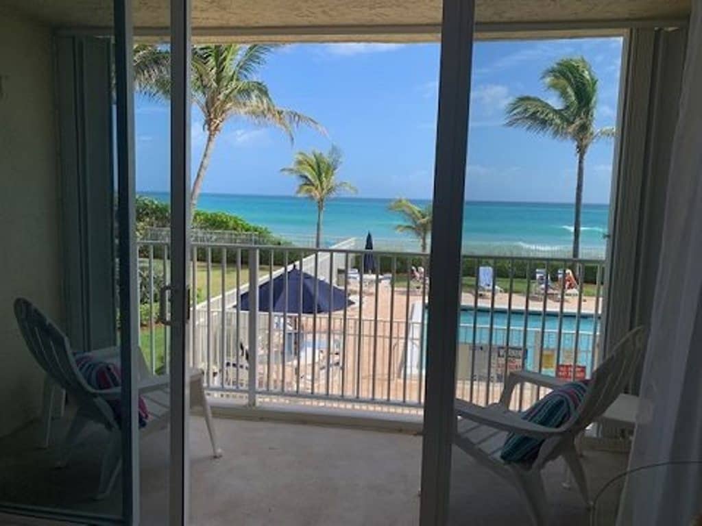 The image captures a view from a balcony, where two white chairs face the ocean. Beyond the balcony railing, turquoise water meets a clear blue sky, with palm trees swaying gently in the breeze. A heated pool is visible below, surrounded by poolside umbrellas.