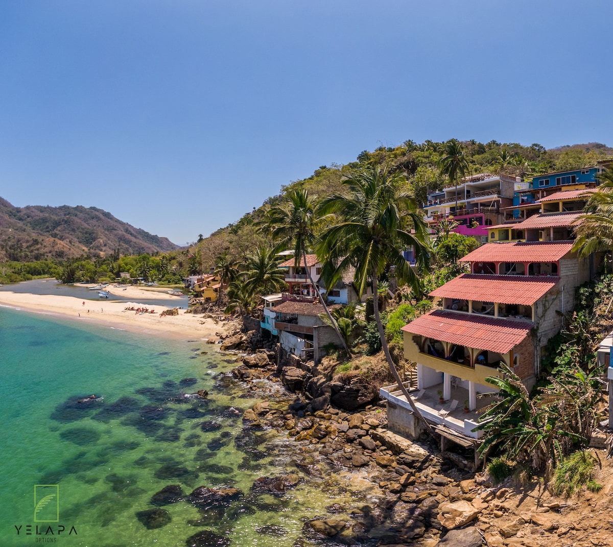 A sandy beach stretches alongside clear turquoise waters, bordered by rocky formations. Colorful structures sit against the lush green hillside, with palm trees swaying gently in the breeze. The scene captures a serene coastal landscape, perfect for nature lovers seeking tranquility.