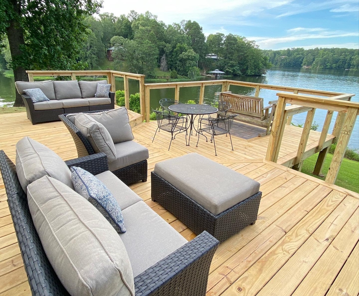 A spacious deck overlooking Lake Jordan features a comfortable seating arrangement with plush gray cushions. A round dining table sits adjacent to a wooden bench, all surrounded by a wooden railing that frames scenic water views and lush greenery.