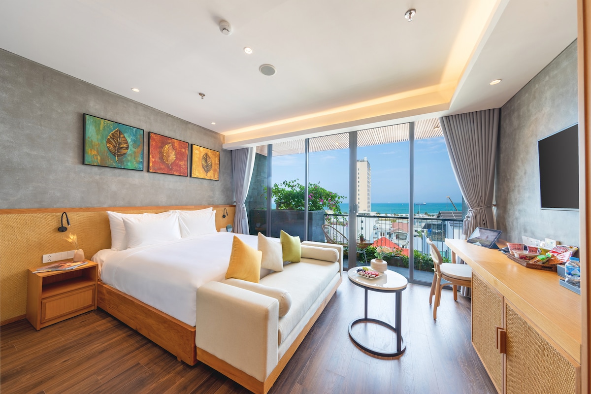 A modern guest room features a spacious bed with decorative cushions, complemented by a seating area. Large glass doors open to a balcony adorned with greenery and ocean views. Artwork with natural themes enhances the stylish decor, and a small table is set nearby.