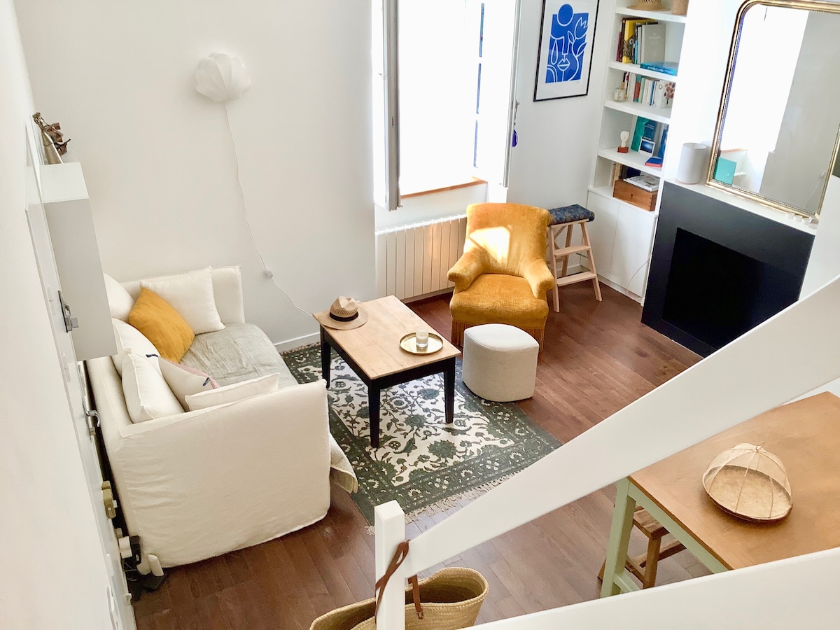 The living area features a white sofa and a yellow accent chair, positioned on a patterned rug. A wooden coffee table is centrally located, alongside a stool. Natural light fills the space through windows, and shelves containing decor and books can be seen in the background.