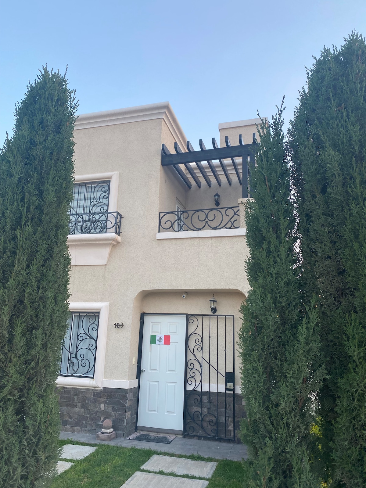 The exterior of the house features a light-colored façade with decorative wrought iron balcony railings. Tall green trees frame the entrance, which is marked by a white door and subtle stone accents. A pergola above the balcony adds architectural interest.
