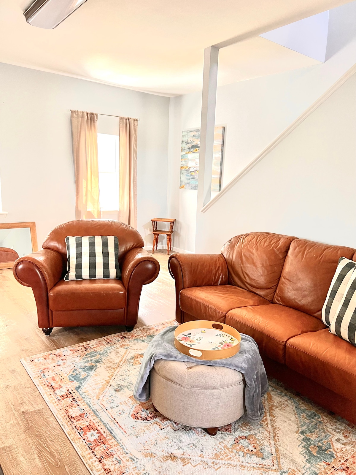 A comfortable living area features a brown leather sofa and an armchair with striped cushions. A round ottoman sits in the center, topped with a decorative tray. Natural light enters through a window dressed with light curtains, enhancing the welcoming ambiance.