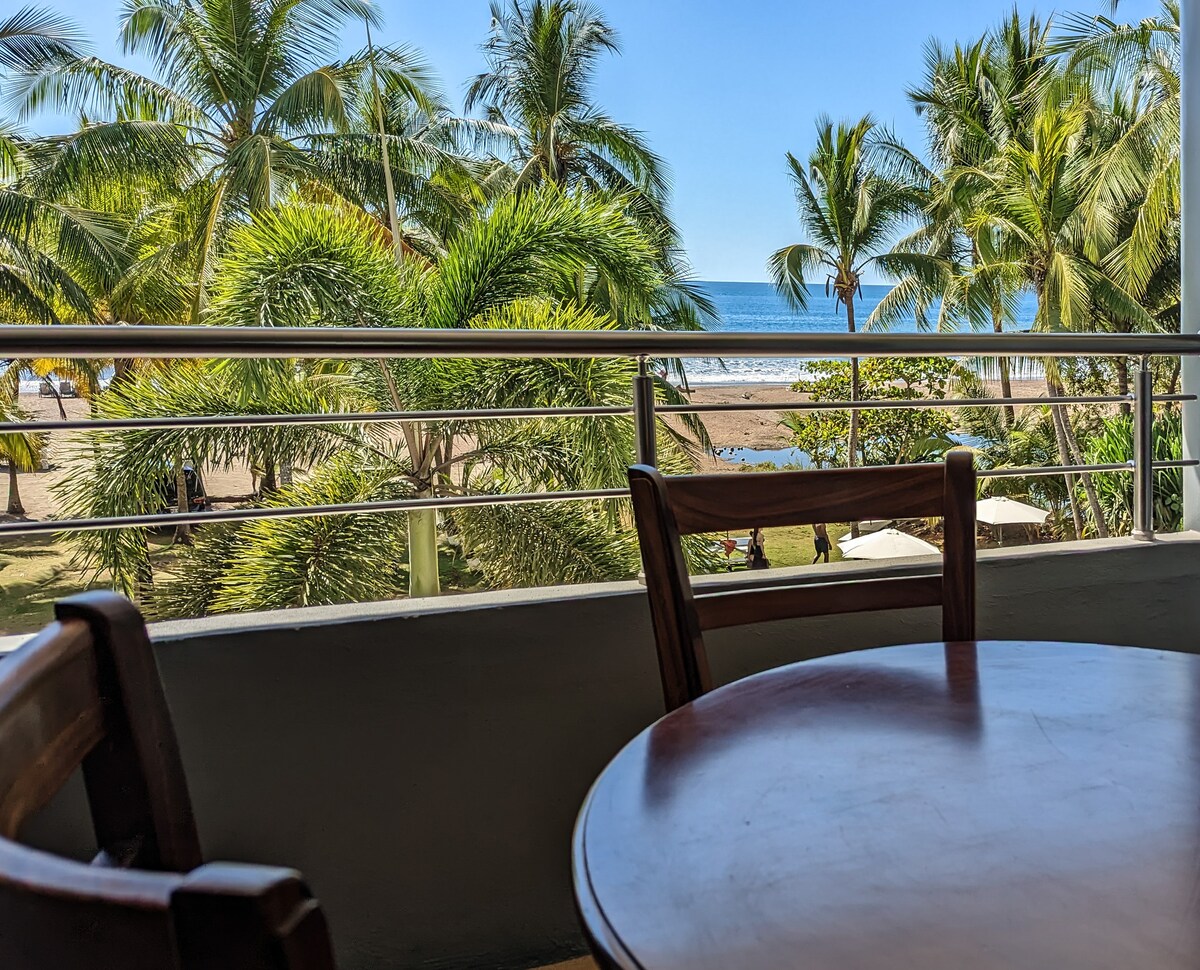 A private balcony view displays lush palm trees framing the Pacific Ocean. A circular wooden table with two chairs is positioned near the balcony's edge, inviting relaxation while enjoying the scenery. The beach is visible beyond the greenery, suggesting proximity to a serene coastal escape.