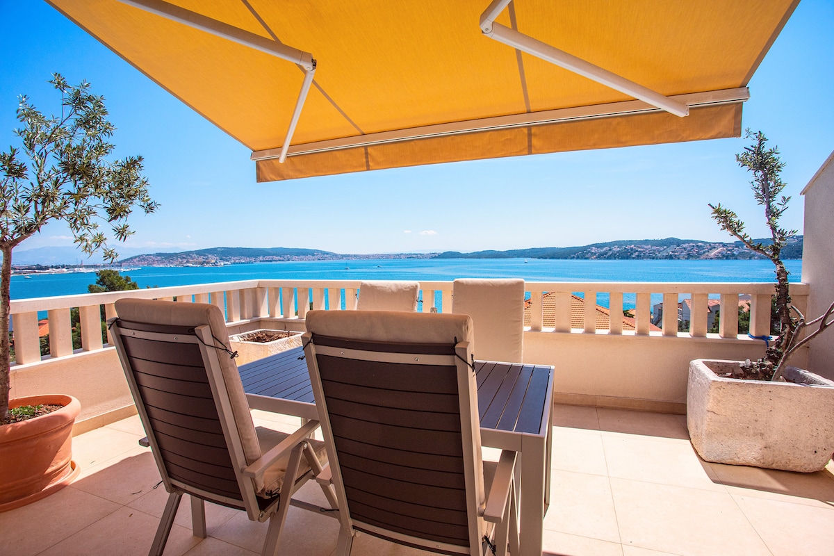 A spacious terrace features a shaded dining area with a table and chairs, providing panoramic views of the water. Potted plants add greenery to the space, while a large awning protects against sunlight, enhancing the relaxed outdoor atmosphere.