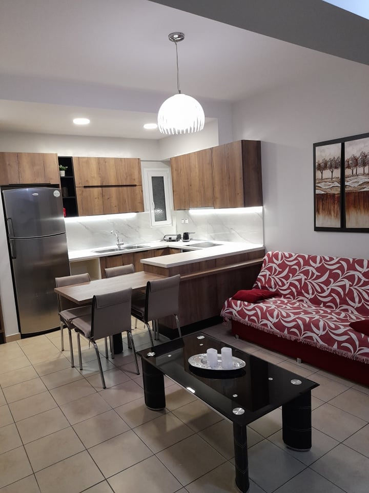 A modern kitchen area features wooden cabinetry and stainless steel appliances, including a refrigerator. A dining table with four chairs is positioned nearby, alongside a red and white patterned sofa. A glass coffee table is centrally located, with decorative candles displayed on top.