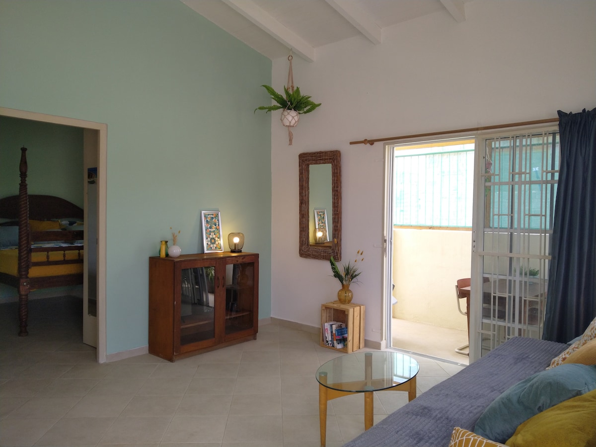 A living area features a comfortable sofa and a round glass table. A wooden cabinet displays decorative items and a mirror reflects natural light. A doorway leads to a bedroom, while another opening gives access to an outdoor area with a small table and chairs.