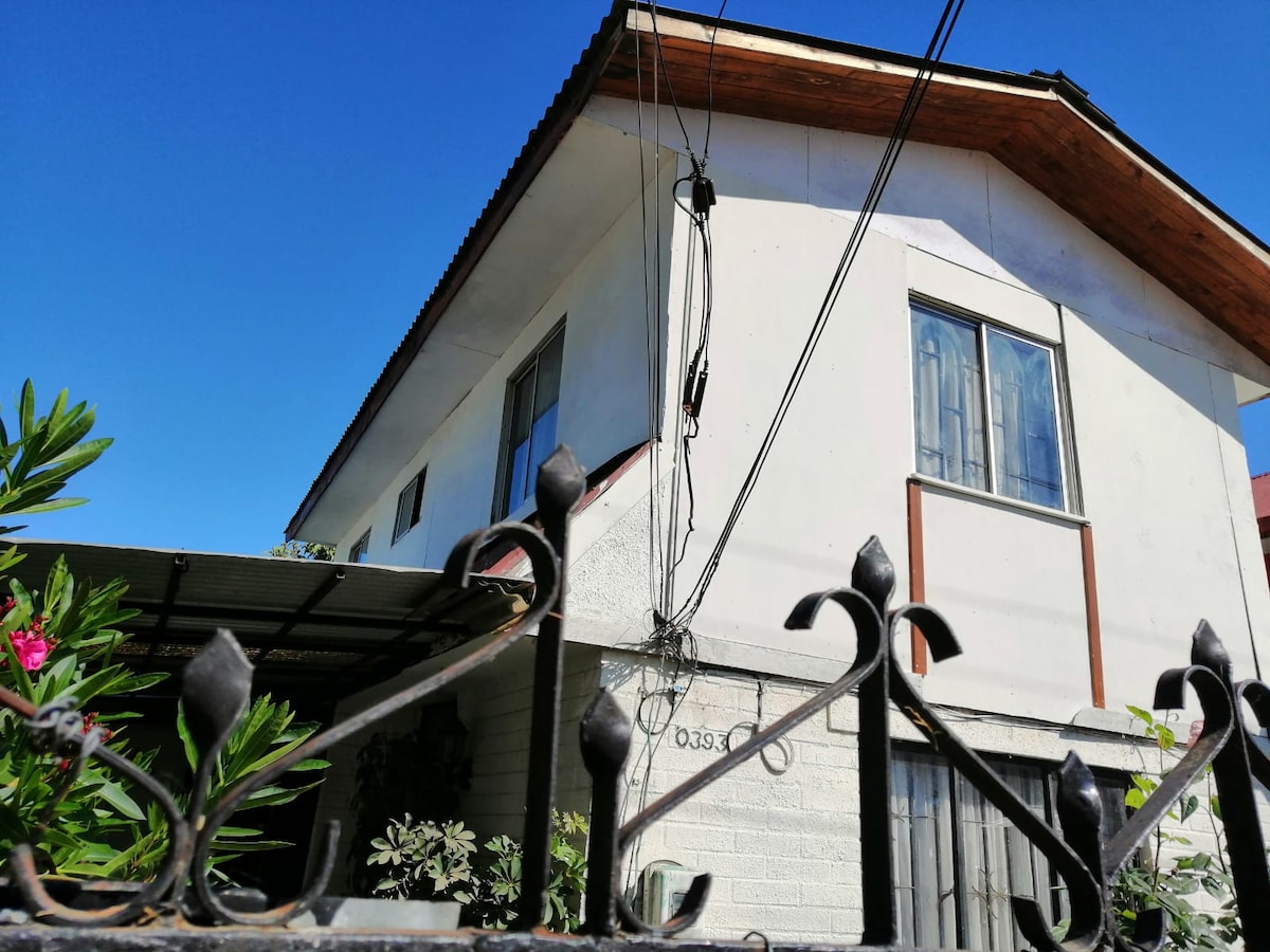 The exterior of the house features a white façade with wood accents and large windows. A pitched roof is visible above, and plants surround the entrance. A decorative wrought-iron gate and fence border the front, providing an inviting approach to the property.