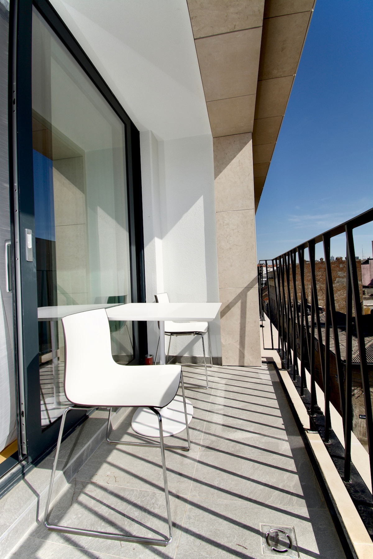A sunny balcony is equipped with a small white table and two modern chairs. The balcony features a railing providing a sense of openness, while the view reveals rooftops and a clear sky, enhancing the outdoor space.