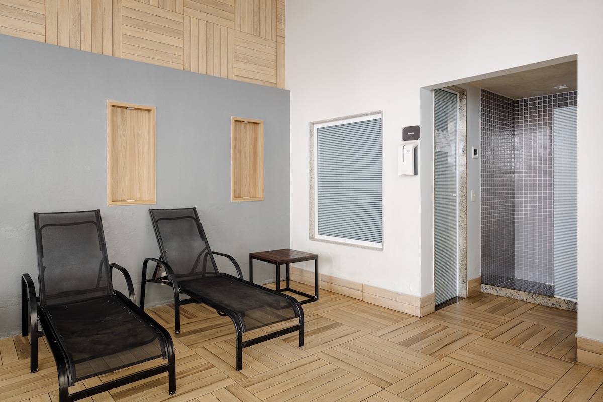 An indoor relaxation area is presented with two lounge chairs positioned beside a wooden table. The warm-toned wooden flooring contrasts with the gray walls, while a tiled shower enclosure is visible through an archway to the right.
