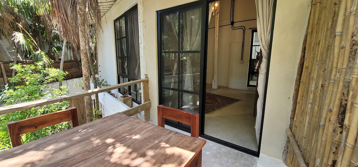 An outdoor dining area features a wooden table and chairs, set against a backdrop of lush greenery. Large glass doors offer a view into the interior space, showcasing an airy atmosphere with soft lighting and minimalistic decor.