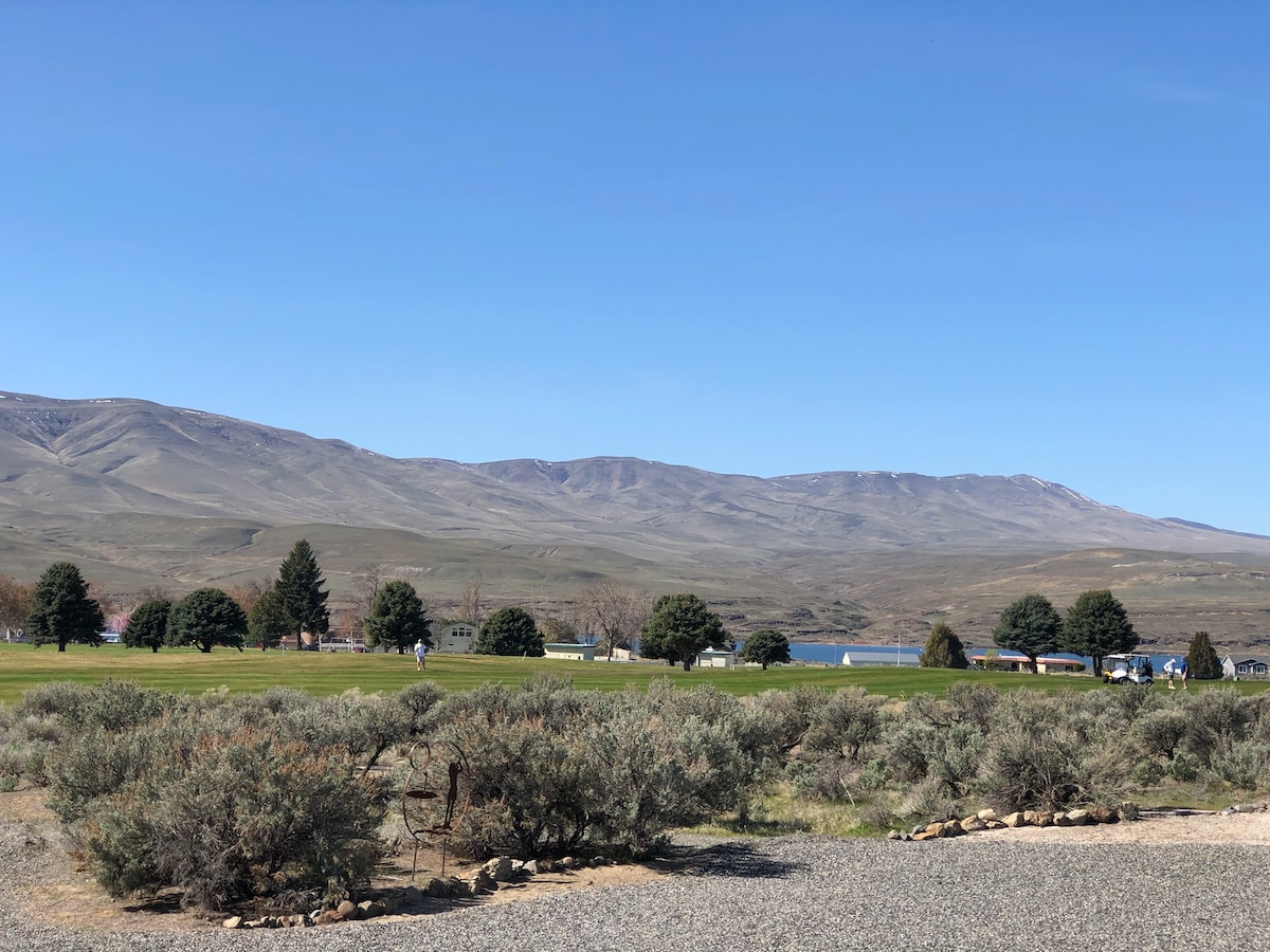 A scenic view showcasing rolling hills and mountains under a clear blue sky. A green landscape with trees and shrubs is visible in the foreground, leading to a tranquil body of water in the distance. The area reflects the natural beauty of the surroundings.