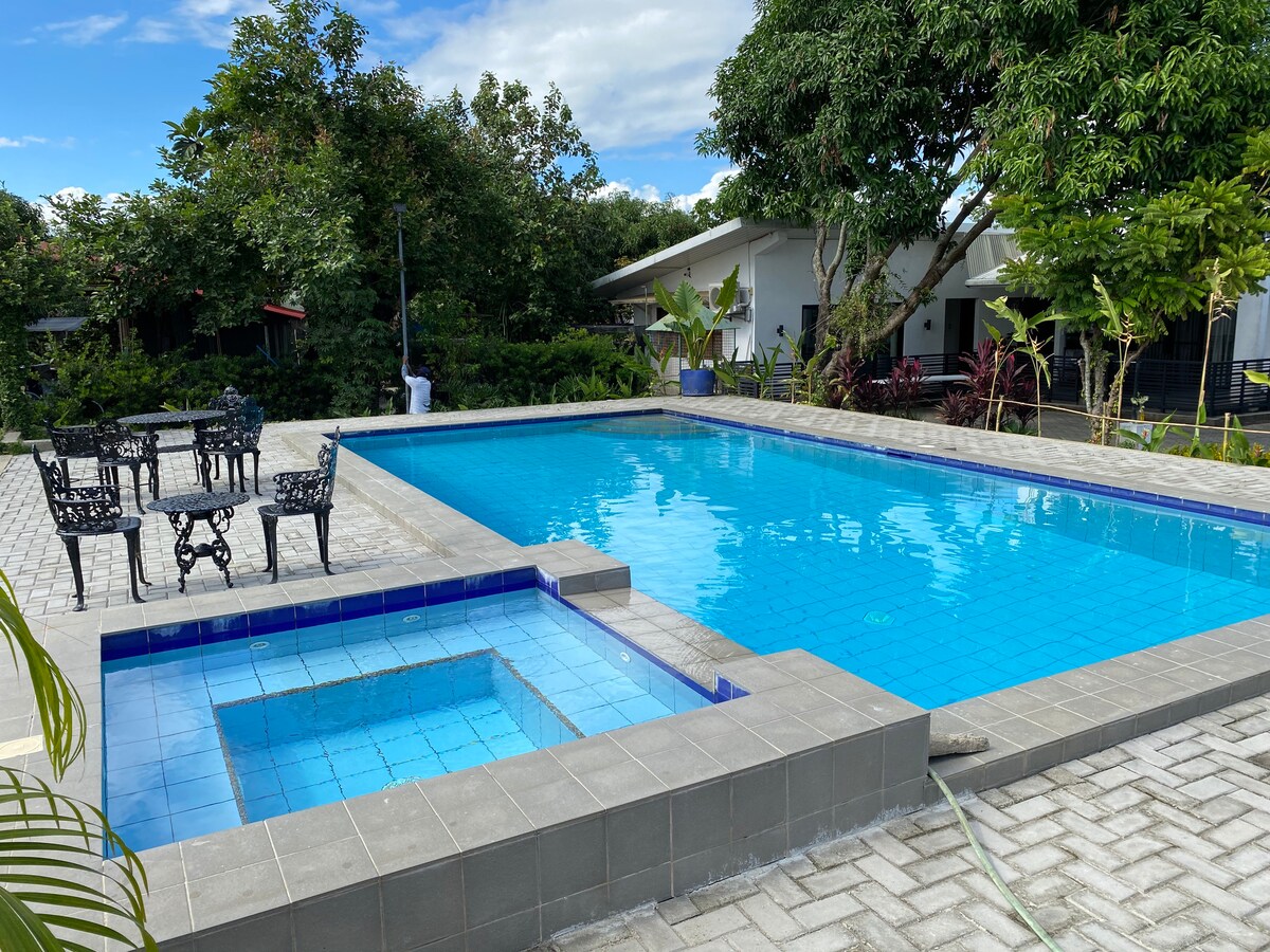 A clear swimming pool is surrounded by spacious paving and lush greenery. A smaller hot tub is positioned adjacent to the pool. Metal chairs and a table are set nearby, offering a spot for relaxation in nature.
