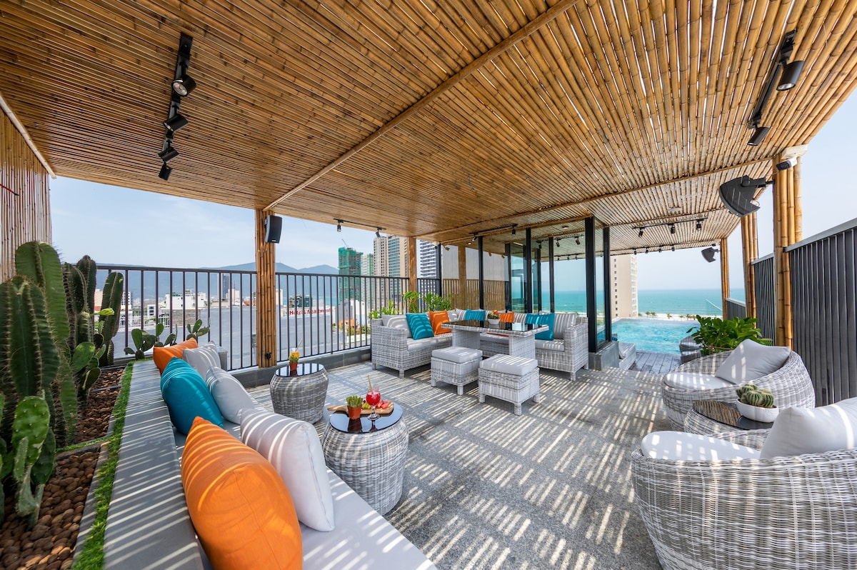 A spacious rooftop terrace is decorated with vibrant cushions on comfortable seating. Natural wooden beams create an inviting overhead structure, while potted cacti and plants enhance the setting. A panoramic view of the ocean is visible in the background, contributing to the serene atmosphere.