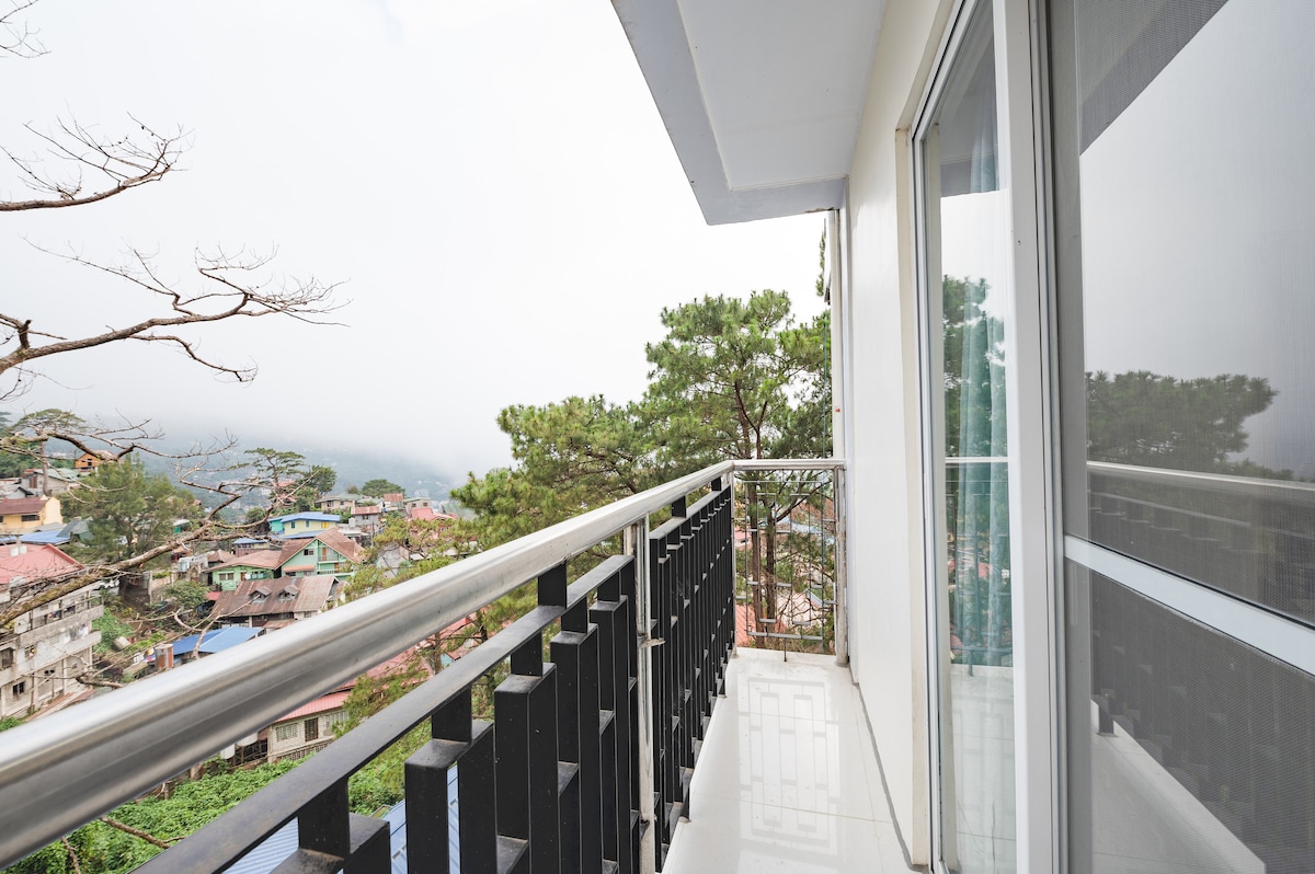 A balcony is observed with a sleek railing overlooking a misty landscape. Nearby trees provide a natural frame, while the distant view reveals a collection of colorful houses nestled among the hills. The space is bright and inviting, featuring smooth flooring.
