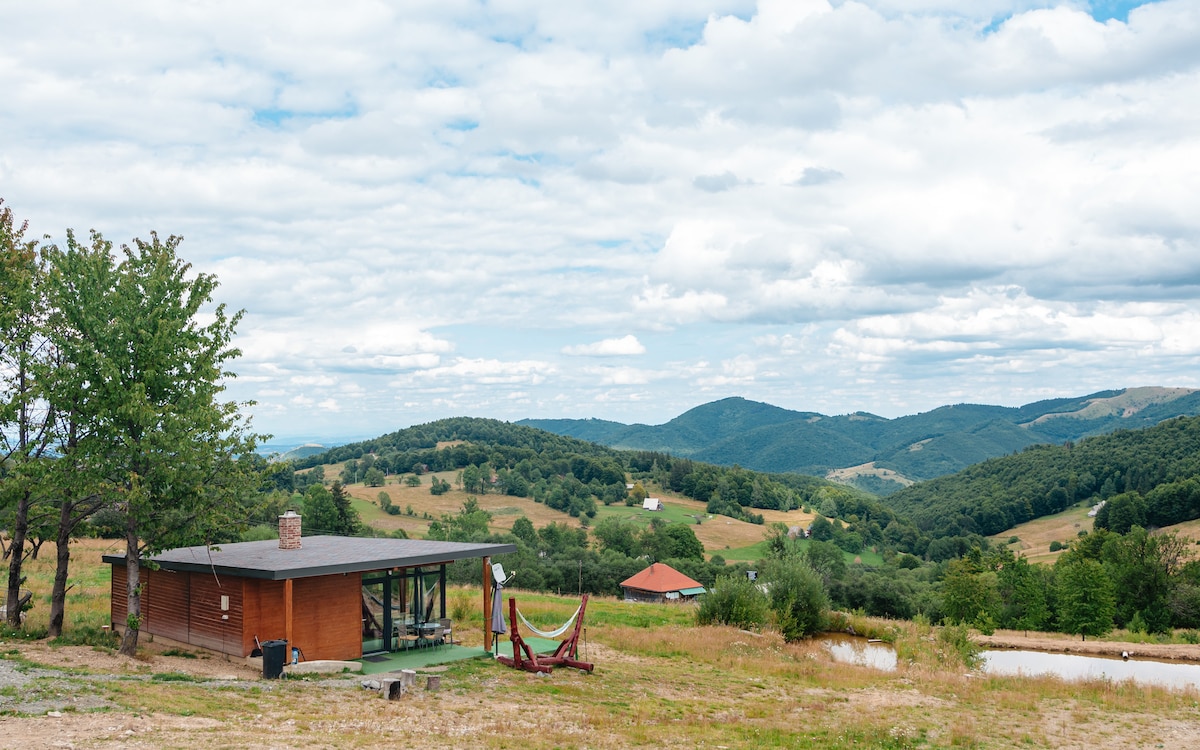 A modern wooden structure is set against a backdrop of rolling hills and expansive skies. A hammock is suspended between two trees nearby, inviting relaxation. The landscape features green fields and distant mountains, providing a serene natural setting.