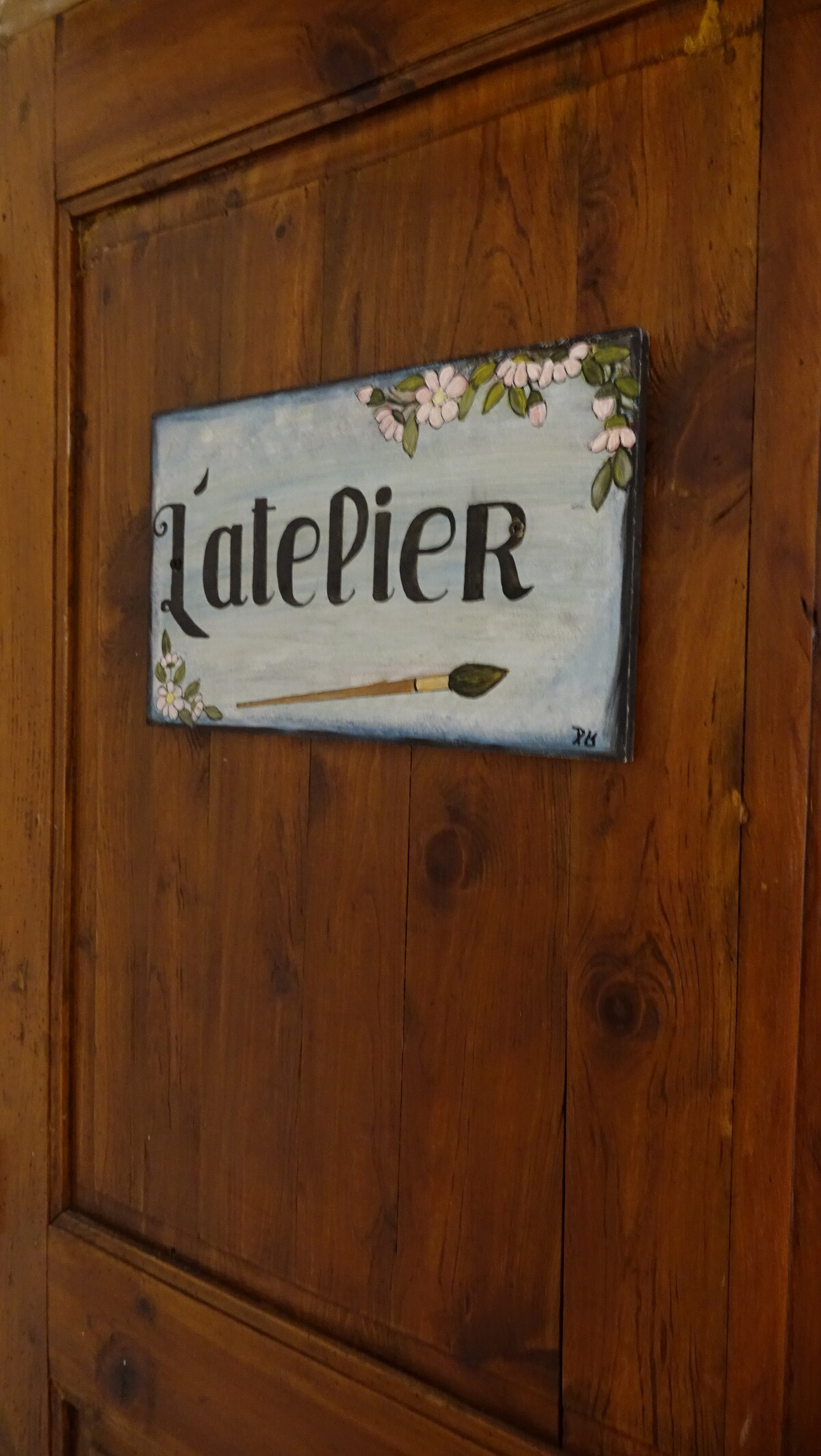 A wooden door features a hand-painted sign reading 'L'atelier', adorned with floral designs and a paintbrush illustration. The rich grain of the wood enhances the rustic charm of the entrance.