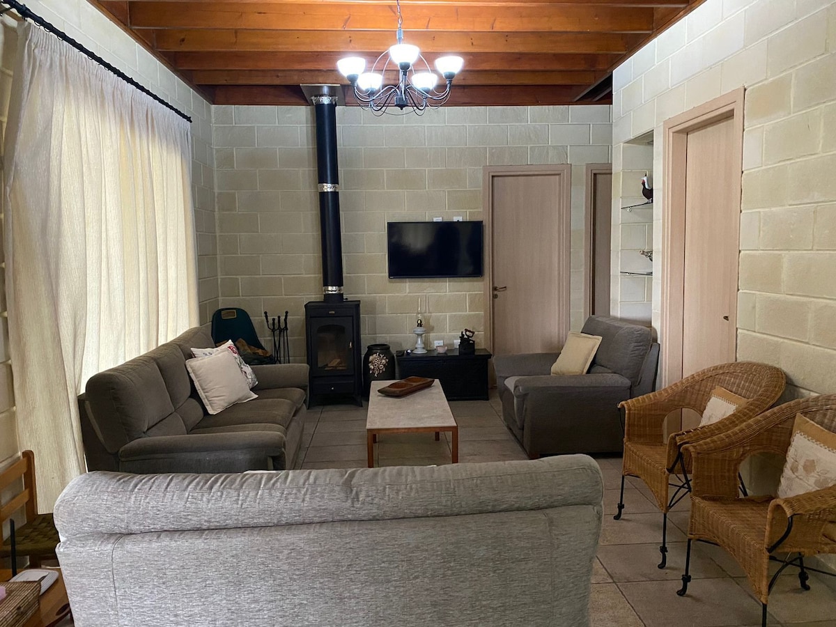 A living area features a mix of comfortable seating options, including a spacious sofa, armchairs, and a central coffee table. A wood stove is positioned against a stone wall, and a flat-screen television is mounted nearby. Soft natural light enters through large curtains.