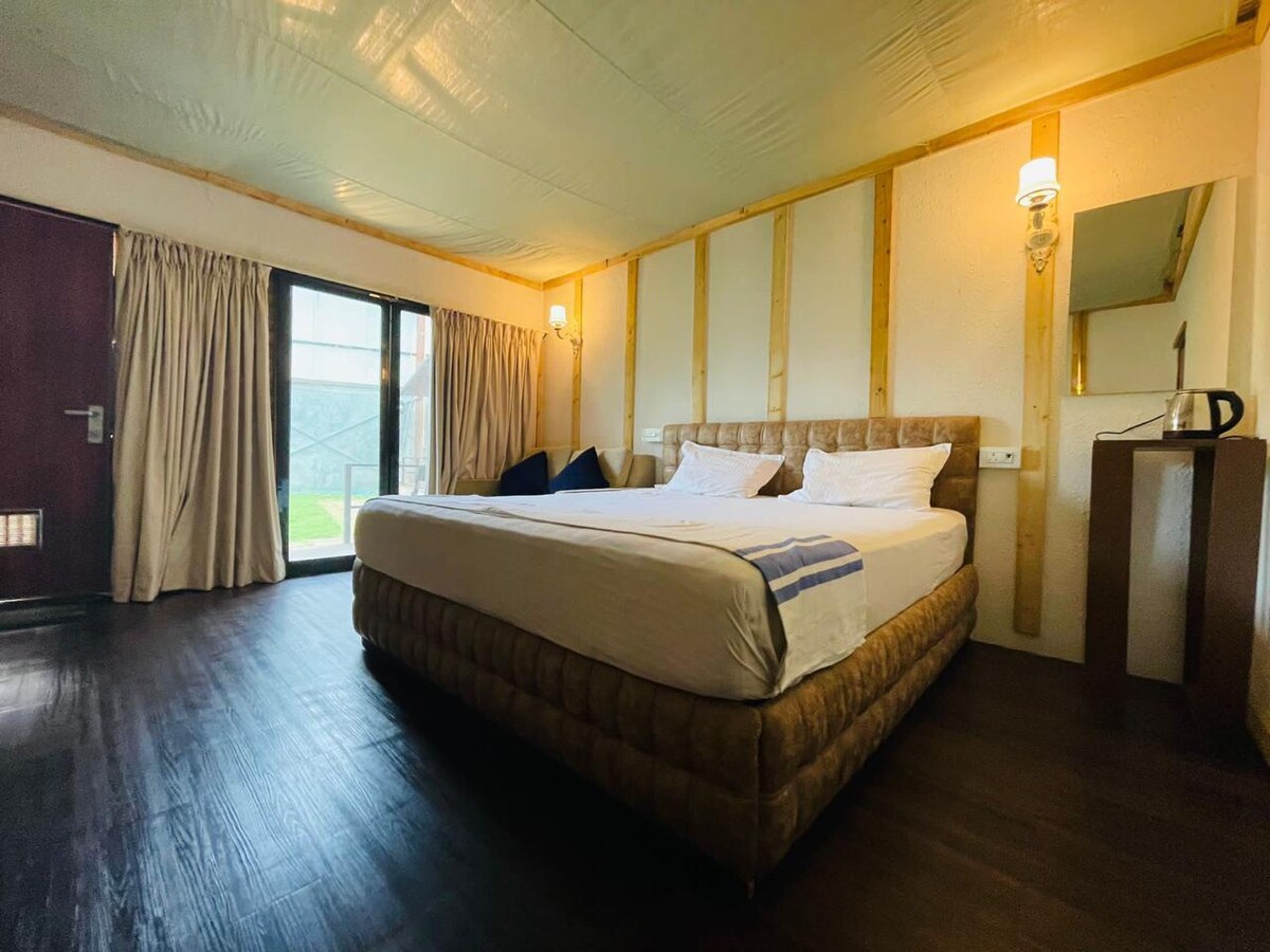 A spacious room features a large, comfortable bed with light bedding against a wooden accent wall. Natural light fills the space from the glass door leading to the outside, complemented by two bedside lamps and a mirror. Curtains frame the door, adding an elegant touch.