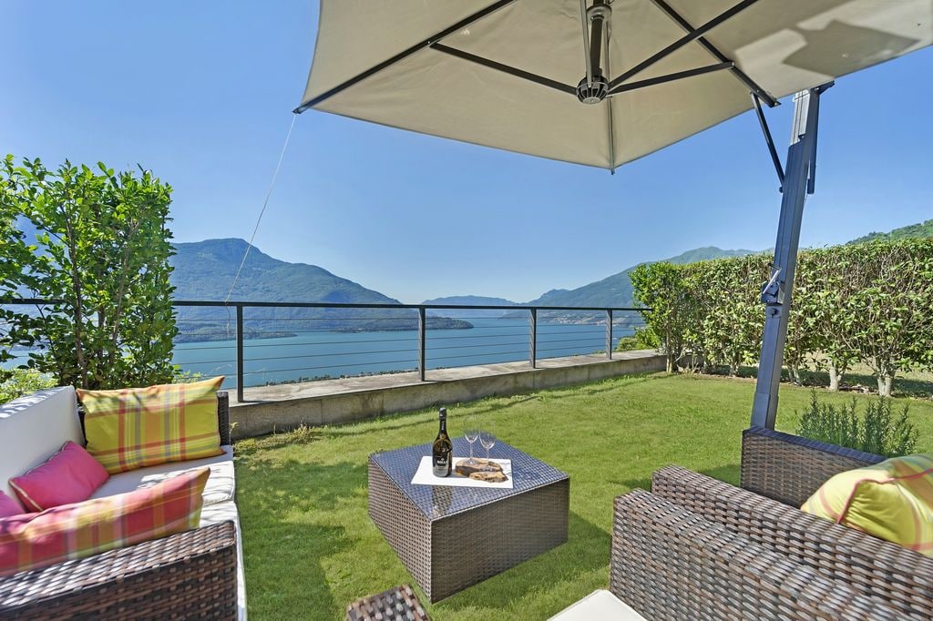 An outdoor terrace features a shaded seating area with a wicker sofa adorned with colorful cushions. A small table is set with two glasses and a bottle, inviting relaxation. The panoramic view of the lake and mountains serves as a stunning backdrop.