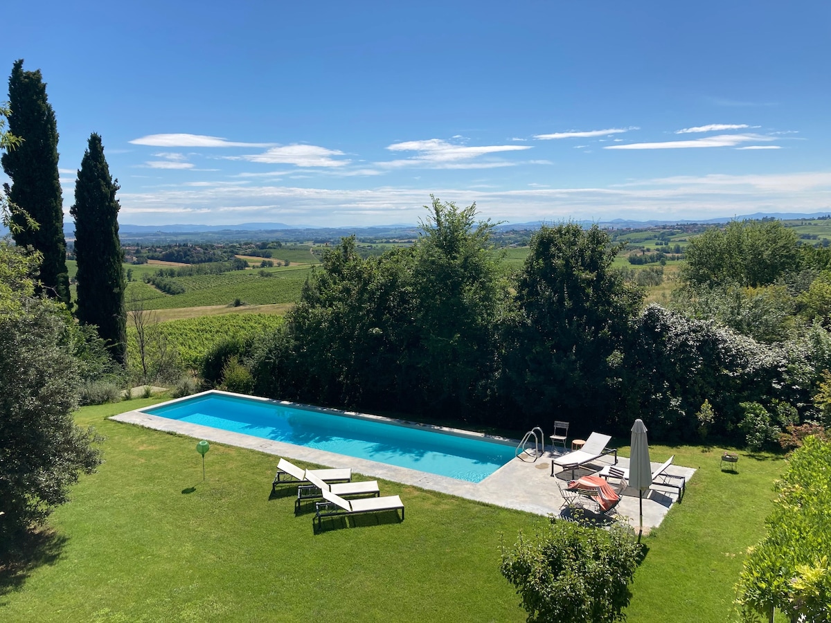 A large, rectangular swimming pool is set within a green lawn, surrounded by tall trees and blooming shrubs. Sun loungers are placed around the pool, providing a space for relaxation. The lush Tuscan countryside stretches into the distance under a clear blue sky.